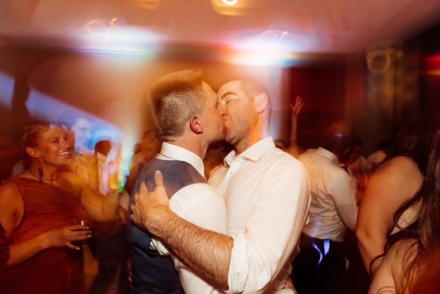Two grooms embrace and kiss on the dance floor while wedding guests smile and cheer around them at their Palm Springs desert resort gay wedding with professional DJ entertainment